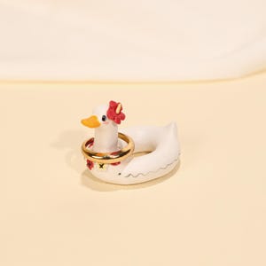 Könnte beinhalten: Ein weißer Keramik-Enten-Ringhalter mit einem goldenen Ring um den Hals. Die Ente hat einen gelben Schnabel, rote Blumenakzente und einen Blumenkranz. Der Ringhalter befindet sich auf einer hellgelben Oberfläche.