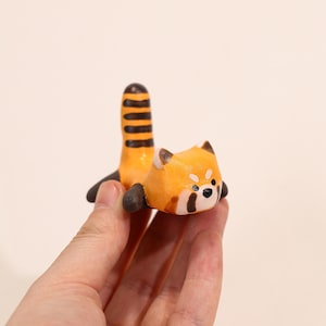 May include: A handcrafted red panda figurine with a distinctive orange and black striped tail. The figurine has a detailed face with black and white accents, and dark brown paws. The overall design is cute and whimsical.