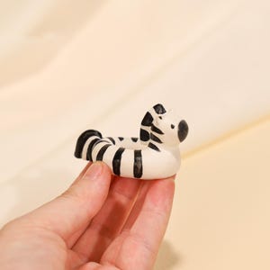 Puede incluir: Pequeña figura de cebra de cerámica, con rayas negras sobre base blanca. La cebra tiene un cuerpo redondeado y una cabeza detallada, con ojos negros y nariz negra. La figura se sostiene en una mano sobre un fondo color crema.