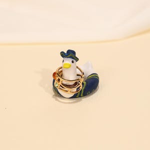 May include: A whimsical ring holder shaped like a duck, adorned with a blue hat and a navy blue and green patterned life preserver. The duck holds a gold ring with colorful gemstones.