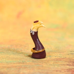 Może przedstawiać: Kapryśna, ręcznie wykonana figurka przypominająca ptaka. Rzeźba ma ciemnofioletowy korpus, żółty dziób i grzebień oraz złoty pierścień wokół podstawy. Oczy są szczegółowe w kolorze białym i czarnym. Tło to mieszanka żółtego i pomarańczowego.