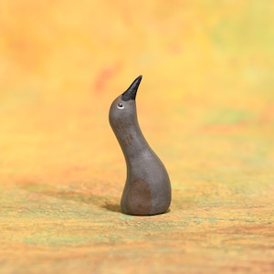Könnte beinhalten: Kleine, handgefertigte Keramik-Vogelfigur. Der Vogel hat einen grauen Körper mit einem dunkelbraunen Fleck und einem schwarzen Schnabel. Sein Kopf ist nach oben geneigt und er hat ein kleines, weißes Auge. Die Figur steht vor einem gelb-grünen Hintergrund.