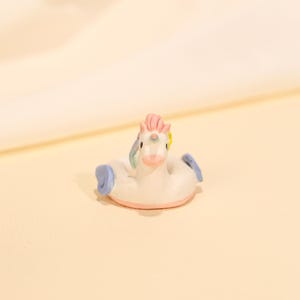 Puede incluir: Un flotador de piscina en miniatura con forma de unicornio. El flotador es blanco con una base rosa y reposabrazos azules. El unicornio tiene una melena rosa, una flor amarilla y una cola de arcoíris. El fondo es de color beige claro.