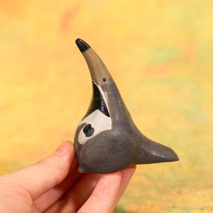 Peut inclure: Figurine d'oiseau en céramique peinte à la main, dans les tons de gris, marron, noir et blanc. L'oiseau a un long bec incurvé et un corps arrondi. La figurine est un objet décoratif.