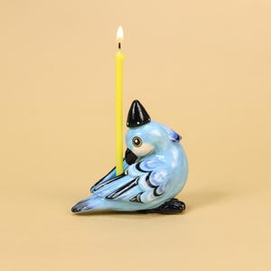 May include: A light blue ceramic bird-shaped candle holder with a black and white hat, holding a lit yellow candle. The bird has black and white wing details and a black beak. The background is a solid tan color.