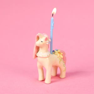 Könnte beinhalten: Ein skurriler Einhorn-Geburtstagskerzenhalter in Creme, Rosa und Gelb. Das Einhorn hat eine hellblaue Kerze mit einer brennenden Flamme. Das Einhorn ist mit Blumen verziert und hat eine rosa Mähne und einen rosa Schweif. Der Hintergrund ist einfarbig rosa.
