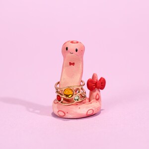 May include: A whimsical, pink ceramic ring holder shaped like a smiling worm with a red bow tie and bow. A gold ring with colorful gemstones rests on the worm's body. The ring holder is on a pink surface.
