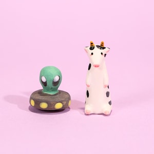 May include: Two ceramic figurines on a pink background. One is a green alien with large white eyes atop a brown base with yellow dots. The other is a white cow with black spots and small yellow horns.