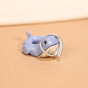 May include: A light blue whale-shaped ring with a silver band. The whale has a white belly, a black eye, and a small fin. The ring is a unique piece of jewellery.