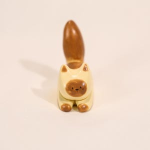 May include: A wooden cat figurine with a cream-colored body and a brown face, tail, and paws. The cat is in a sitting position with its tail pointing upwards. The figurine has a simple, cartoonish design.
