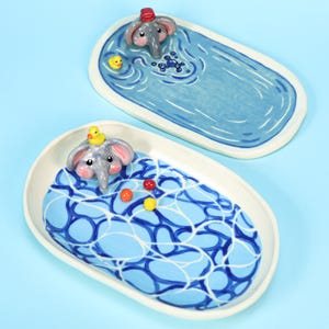 May include: Two ceramic elephant-themed trinket dishes. One features an elephant in a bathtub with a rubber ducky. The other has an elephant under a blue blanket with colorful balls. Both dishes have a white rim and are on a blue background.