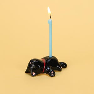 Puede incluir: Un portavelas de cerámica negra con forma de pantera con una raya roja y una vela azul claro con una llama encendida. El portavelas está sobre un fondo amarillo.