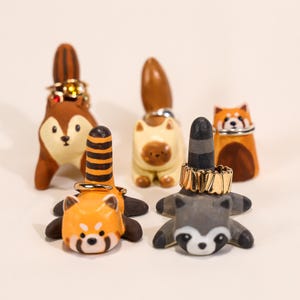 May include: A selection of animal-shaped ring holders. The holders feature a squirrel, a cat, a red panda, and a raccoon. Each holder has a post for rings. The squirrel is brown and cream, the cat is cream, and the red panda is orange and black. The raccoon is grey.