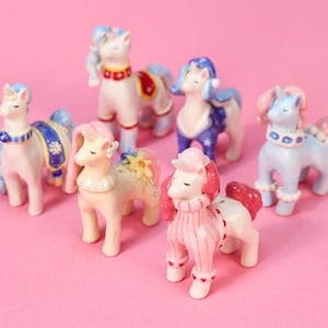 May include: A collection of ceramic pony figurines in various pastel colors, including pink, blue, and cream. Each pony has unique decorative elements like floral accents, patterned saddles, and ruffled collars. The ponies are arranged on a pink background.
