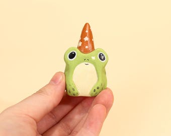 Op de afbeelding: Een kleine, groene kikkerfiguur met een witte buik en grote zwarte ogen. De kikker draagt een puntige oranje feesthoed met witte steraccenten. Het beeldje wordt in een hand gehouden tegen een lichtgele achtergrond.