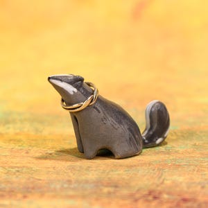 Könnte beinhalten: Grau-weißes Keramik-Dachs-Figürchen mit einem silbernen Ring um den Hals. Der Dachs sitzt, der Schwanz ist gekrümmt. Der Hintergrund ist eine Mischung aus Gelb und Orange.