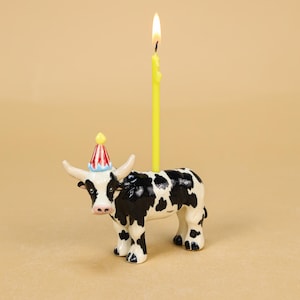 Peut inclure: Figurine de vache en céramique avec des taches noires et blanches, portant un chapeau de fête et tenant une bougie jaune allumée. La vache a un nez rose et se tient sur une surface beige. La bougie mesure environ 15 cm de haut.
