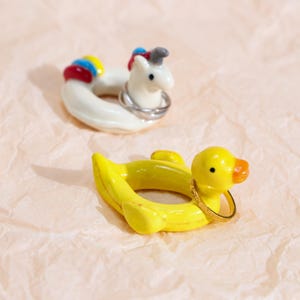 Peut inclure: Deux porte-bagues fantaisistes : une licorne blanche avec une crinière arc-en-ciel et un canard jaune avec un bec orange. Les deux sont conçus pour contenir des bagues, ajoutant une touche ludique à toute collection de bijoux. Sur fond rose clair.