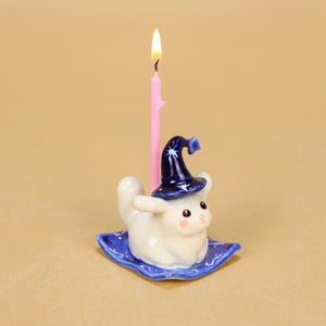 May include: A whimsical birthday candle holder featuring a white ceramic bunny wearing a dark blue wizard hat with white stars. The bunny sits on a blue square base with a wavy design. A pink candle with a lit flame is in the center.