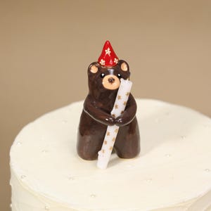 May include: A brown ceramic bear figurine wearing a red party hat with white flower patterns. The bear is holding a white candle decorated with small gold stars. The figurine is positioned on top of a white frosted cake with small white sprinkles.