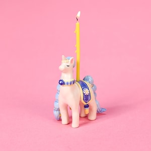 May include: A whimsical unicorn-shaped birthday candle holder. The ceramic figurine is light pink with blue accents on the mane, tail, and decorative saddle. A yellow candle is lit and placed in the center of the unicorn's back. The background is a solid pink.