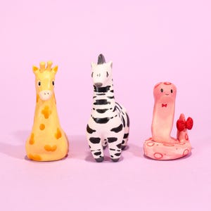 Può includere: Tre figurine di animali stravaganti: una giraffa con macchie arancioni, una zebra con strisce nere e un serpente rosa con un fiocco rosso. Le figurine sono ambientate su uno sfondo rosa.