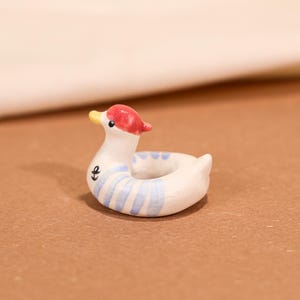Op de afbeelding: Een keramische eendvormige zwemband met een rode pet en gele snavel. Het witte lichaam heeft blauwe strepen en een ankersymbool. Dit decoratieve item is waarschijnlijk een miniatuurbeeldje of ornament.