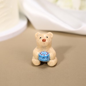 Könnte beinhalten: Eine kleine, glänzende, beige Keramikbärenfigur in sitzender Position. Der Bär hat dunkle Augen, eine rotbraune Nase und hält einen hellblauen zylindrischen Gegenstand, der mit einem Muster aus kleinen roten Punkten und blauen Blumen verziert ist.