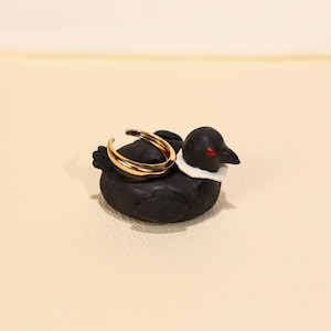 Könnte beinhalten: Ein goldfarbener Ring liegt auf einer kleinen, schwarzen Entenfigur mit weißem Kragen und roten Augen. Der Ring hat ein gebogenes Design. Die Figur befindet sich auf einer cremefarbenen Oberfläche.
