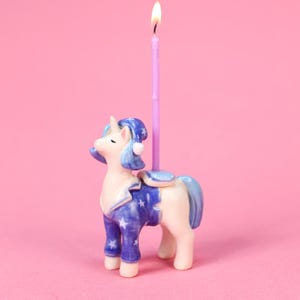 May include: A ceramic unicorn birthday candle holder with a lit purple candle. The unicorn is wearing a blue nightcap and pajamas with a star pattern. The background is a solid pink.