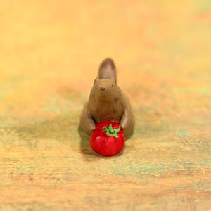 May include: A brown squirrel figurine holding a bright red tomato with green leaves. The squirrel is in a seated position, with the tomato held in its paws. The figurine is set against a blurred, colorful background.