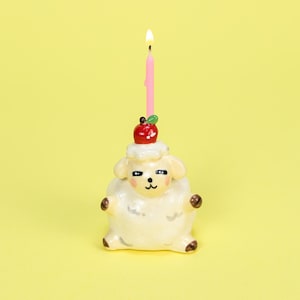 May include: A whimsical, cream-colored sheep-shaped candle holder with a red apple and green leaf on top. A lit pink candle is inserted into the apple. The candle holder has brown accents and is set against a yellow background.