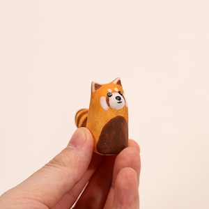 May include: A hand-painted wooden red panda figurine. The figurine features a brown body, orange head and ears, and a striped tail. The red panda has a white face with black eyes and a black nose. The figurine is held in a person's hand.
