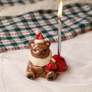 Peut inclure: Figurine d'ours en céramique marron portant un bonnet de Père Noël et un col blanc, tenant un sac rouge. Une bougie argentée allumée est positionnée derrière l'ours. La figurine est sur une surface blanche avec un motif à carreaux vert et blanc en arrière-plan.