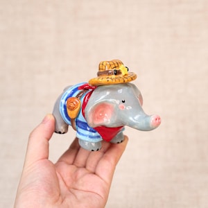 May include: A gray ceramic elephant figurine wearing a blue and white striped shirt, a red bandana, and a yellow hat. The elephant has a trunk, pink ears, and a small saddlebag. A charming decorative item.