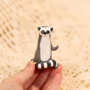 May include: A ceramic lemur figurine with a black and white striped tail. The lemur has a gray body, white chest, and black markings around its eyes. The figurine is small and detailed, with a hand-painted appearance.
