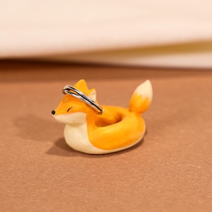 Peut inclure: Un porte-bague décoratif en forme de renard. Le renard est orange et blanc, avec un anneau argenté posé sur sa tête. Le porte-bague est conçu pour contenir des bagues ou de petits bijoux. Le renard est en céramique ou dans un matériau similaire.