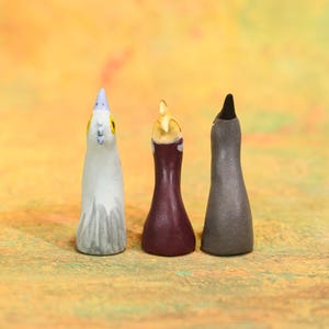 Könnte beinhalten: Drei kleine, handbemalte Keramikfiguren. Die erste ist weiß mit blauem und gelbem Kopf, die zweite ist weinrot mit goldener Spitze und die dritte ist grau mit schwarzer Spitze. Sie sind auf einer strukturierten, gelb-grünen Oberfläche angeordnet.