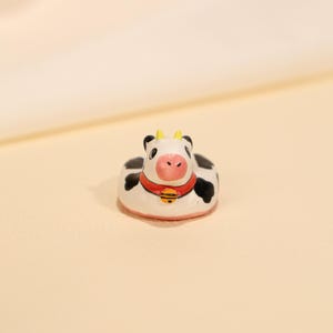 May include: A small, ceramic cow figurine with a white base, black spots, and a pink nose. It has a red collar with a gold bell. The cow has yellow horns and is in a seated position. The figurine is a decorative item.