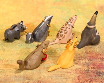 May include: A selection of six ceramic animal ring holders. The animals are a grey elephant, a badger, a spotted seal, a dark brown animal, a brown beaver with a red ball, and a yellow dog. Each animal holds a ring. The ring holders are in various colours.