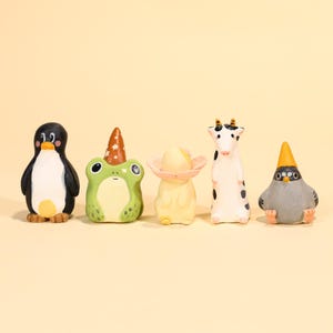 Puede incluir: Un conjunto de cinco figuras de cerámica caprichosas. El conjunto incluye un pingüino, una rana con un gorro de fiesta, una figura con forma de hongo, una vaca y un pájaro con un sombrero amarillo. Cada figura está pintada con colores brillantes y detalles.