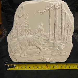 May include: White ceramic wall plaque with a raised design of a wolf howling at the moon in a forest setting. The plaque is approximately 10 inches wide.
