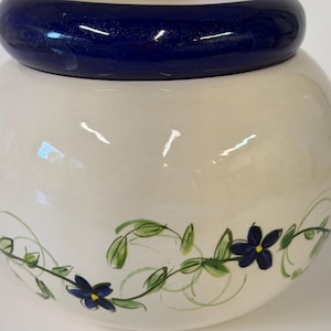 May include: A round ceramic jar with a glossy cream-colored body and a thick, dark blue rim. The lower portion of the jar features a hand-painted design of green vines with small blue flowers that have yellow centers.