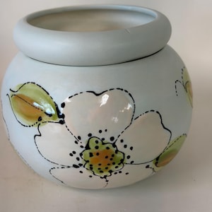 May include: A round, light blue ceramic jar with a thick, rounded rim. The exterior features a hand-painted white flower design with black dotted outlines, green leaves, and a yellow center. The surface has a smooth, glossy finish throughout.