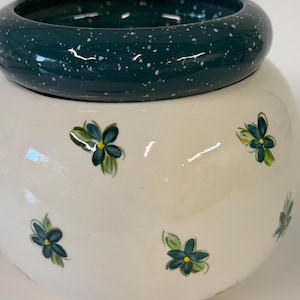 May include: A round ceramic bowl with a cream-colored base featuring small hand-painted dark teal flowers with yellow centers. The rim is a thick, glossy dark teal band with white speckles, creating a speckled finish on the top edge.
