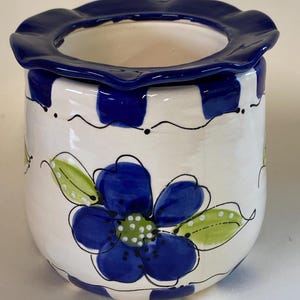 May include: A ceramic flower pot with a white base, adorned with a large blue flower and green leaves. The pot features a blue ruffled rim and blue square patterns around the top. Black wavy lines and dots add detail.