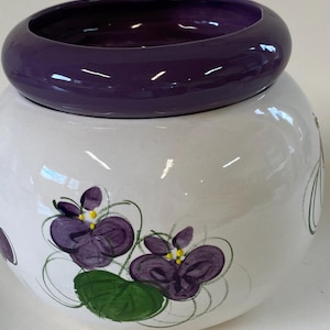 May include: A white ceramic jar with a round, deep purple lid. The jar is decorated with hand-painted purple violets and green leaves. The jar has a round shape.