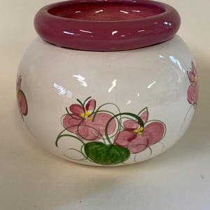 May include: A white ceramic jar with a round, dark pink rim. The jar is decorated with pink floral designs with green leaves and yellow centers. The jar has a rounded shape.