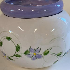 May include: A white ceramic bowl with a lavender rim and floral design. The bowl features hand-painted lavender flowers with yellow centers and green leaves. The design is on a white, round bowl.