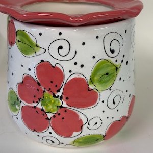 May include: A white ceramic wax warmer featuring a hand-painted floral pattern with red petals, green leaves, and black dotted accents. The top is a removable red scalloped dish, and the cylindrical base is decorated with black swirling line details.